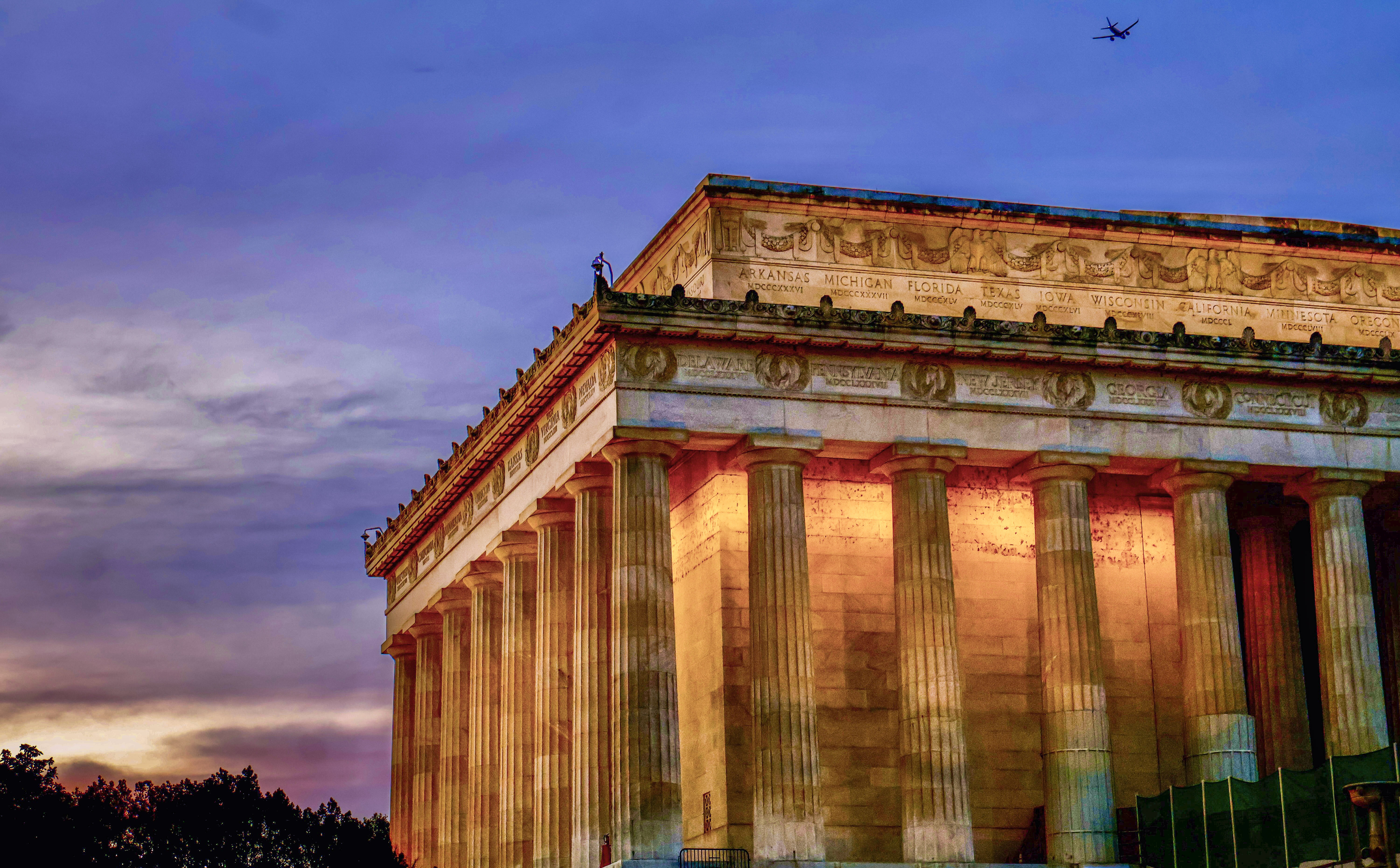 A low-angle view of the Lincoln Memorial at last light, a plane crossing a deep mauve sky.